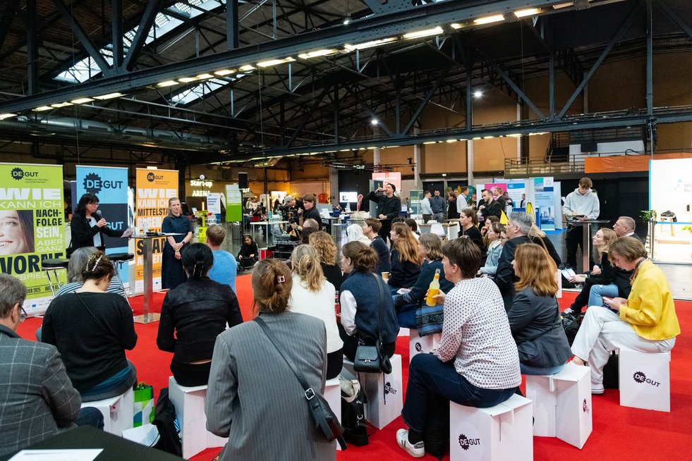 Zahlreiche Gäste sitzen auf Hockern im Bereich Marktplatz auf der Messe deGUT, es wird von den Angeboten des BPW auf der Bühne erzählt und eine Gründerin spricht von ihren Erfahrungen.