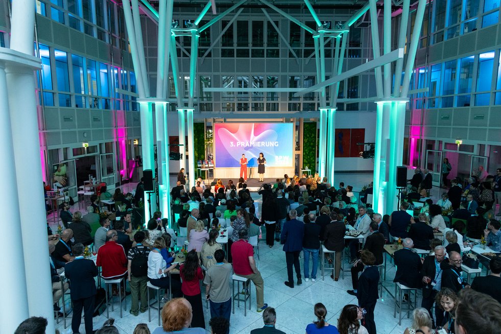 Blick in die Veranstaltungshalle während der dritten Prämierung des Businessplan-Wettbewerb Berlin-Brandenburg (BPW). Zahlreiche Gäste verfolgen das Bühnenprogramm mit zwei Moderatorinnen vor einer großen LED-Wand.