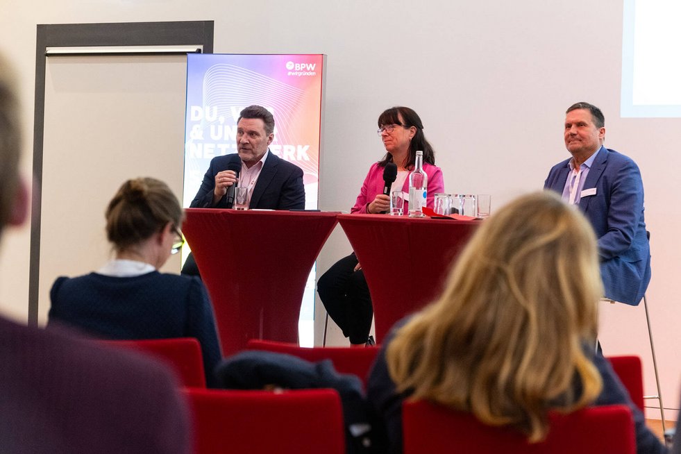 Drei Personen sitzen an Stehtischen auf einer Bühne und unterhalten sich