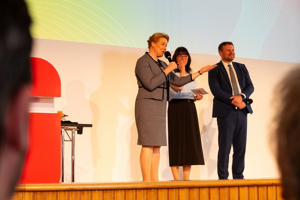 Drei Personen sind auf einer Bühne, eine Frau hält ein Mikrofon und spricht zum Publikum