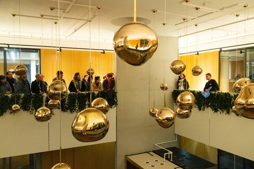 Menschen stehen auf einer Galerie und blicken in einen Innenraum mit hängenden kugelförmigen Lampen.