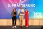 Verleihung Publikumspreis an das Team StemGel, verliehen von Brandenburgs Wirtschaftsministerin Martina Klement Drei Personen stehen auf der Bühne bei der dritten Prämierung des Businessplan-Wettbewerb Berlin-Brandenburg (BPW). Der Gewinner des Publikumspreises präsentiert Urkunde und Trophäe vor einer großen LED-Wand.
