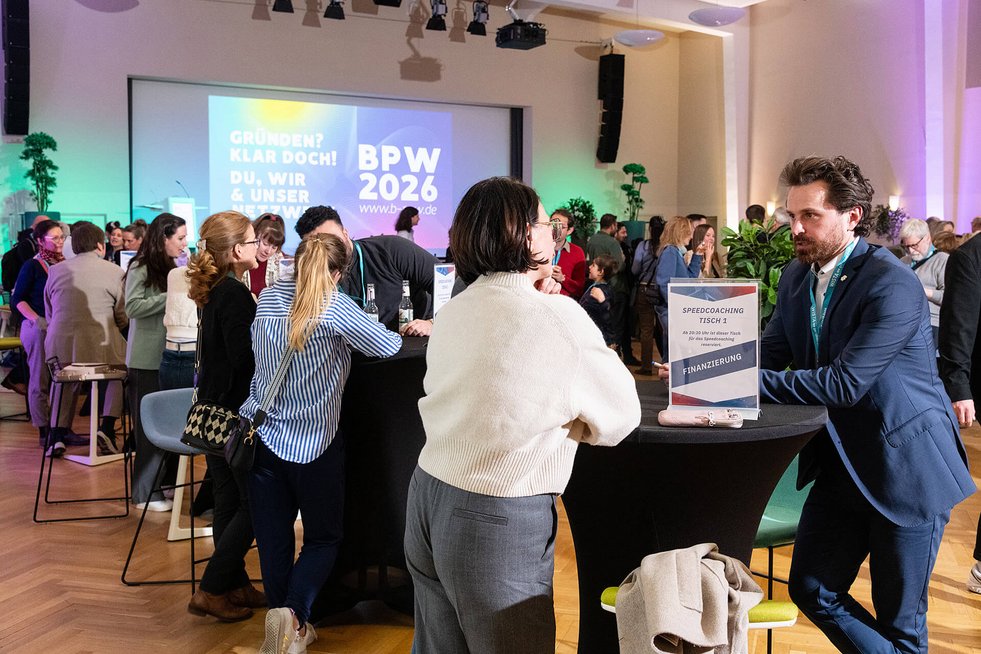 Menschen stehen an Stehtischen und führen Gespräche bei einer Veranstaltung, im Hintergrund eine große Präsentationsfläche.