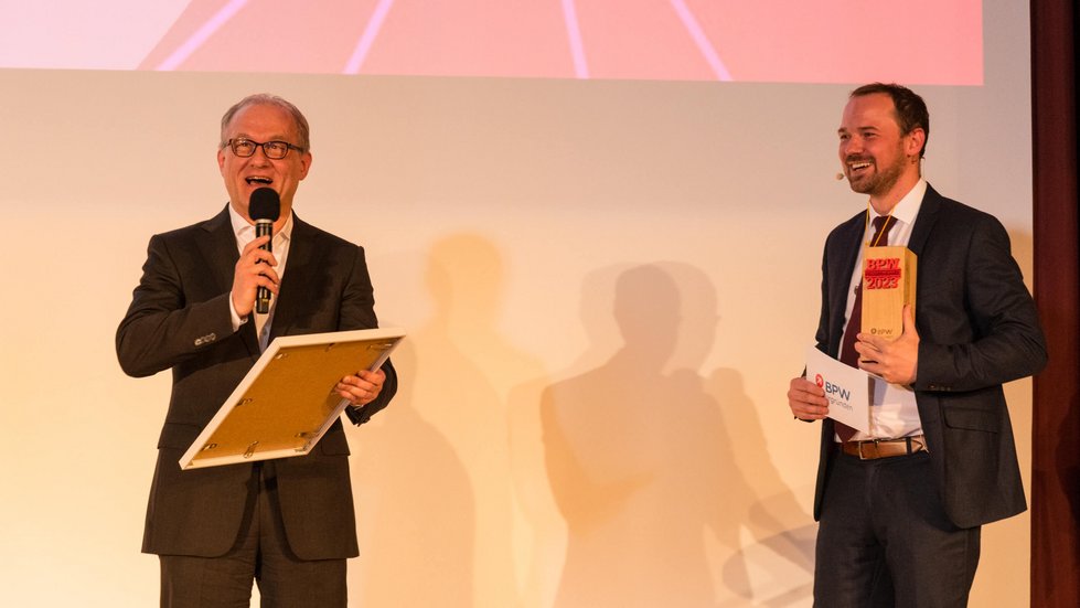 Laudator verkündet ein Siegerteam und hält die Urkunde in der Hand, der Moderator hält einen Pokal