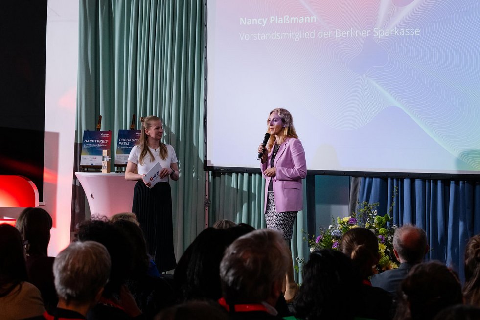 Zwei Frauen stehen auf einer Bühne, eine spricht ins Mikrofon vor Publikum.