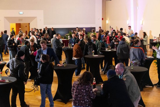 Viele Menschen stehen in einem Veranstaltungssaal und unterhalten sich, während auf einer Bühne eine Präsentation stattfindet.