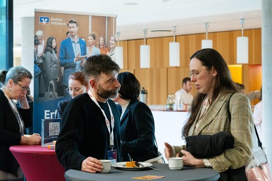 Zwei Konferenzteilnehmende im Gespräch an einem Stehtisch mit Kaffee und Kuchen.