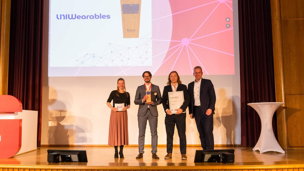 Preisverleihung mit Urkunde und Pokal. Vier Personen stehen für ein Gruppenfoto zusammen.