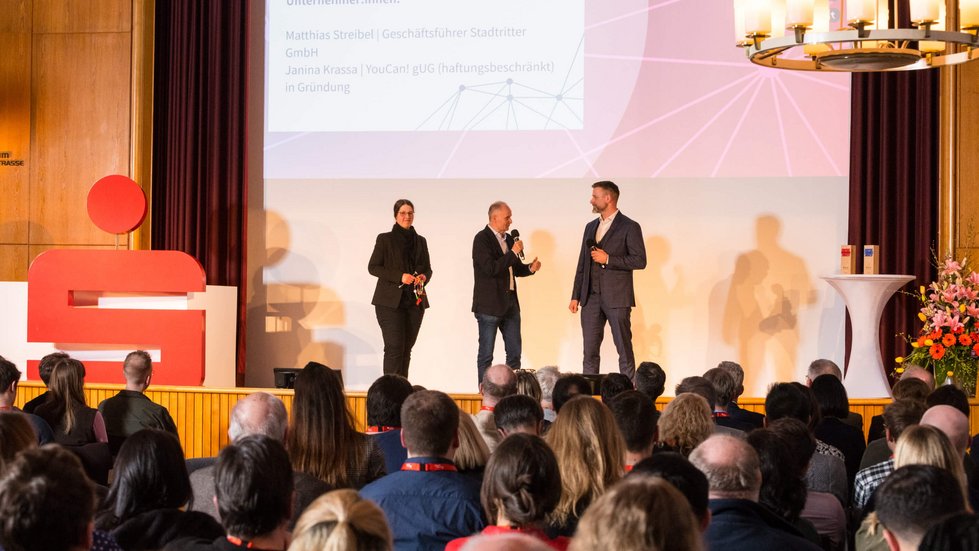 Drei Personen sprechen vor Publikum auf einer Bühne