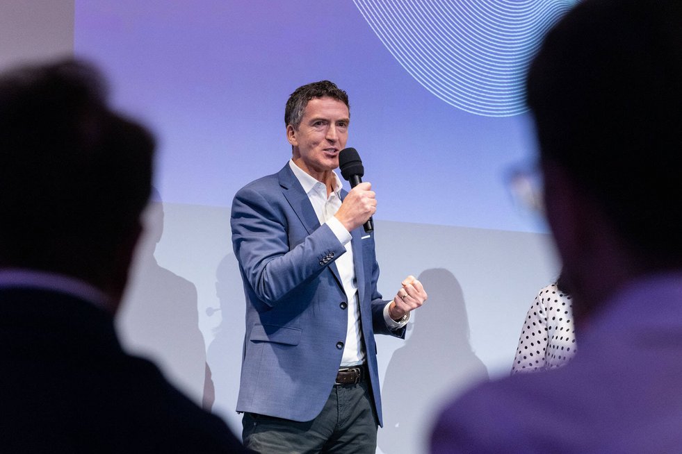 Ein Mann spricht mit einem Mikrofon auf einer Bühne vor Publikum. Er trägt ein Sakko und steht vor einer projizierten Hintergrundgrafik.