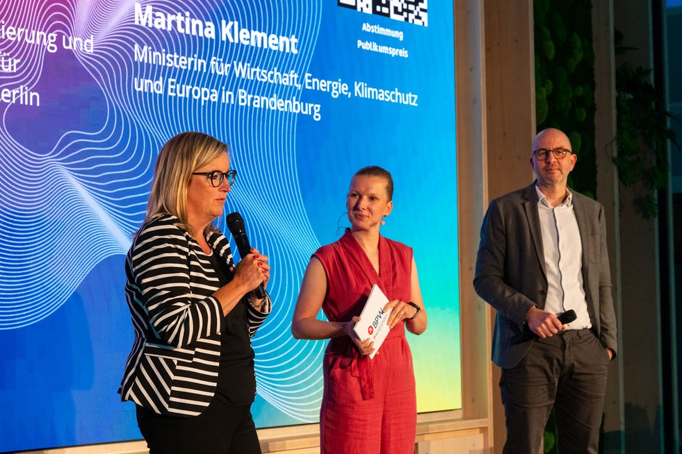 Drei Personen stehen auf der Bühne der BPW-Prämierung. Martina Klement, Ministerin für Wirtschaft, Energie, Klimaschutz und Europa des Landes Brandenburg, spricht ins Mikrofon, während zwei Moderierende daneben stehen.