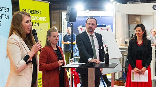 Drei Personen stehen auf einer Bühne, eine Moderatorin spricht zum Publikum
