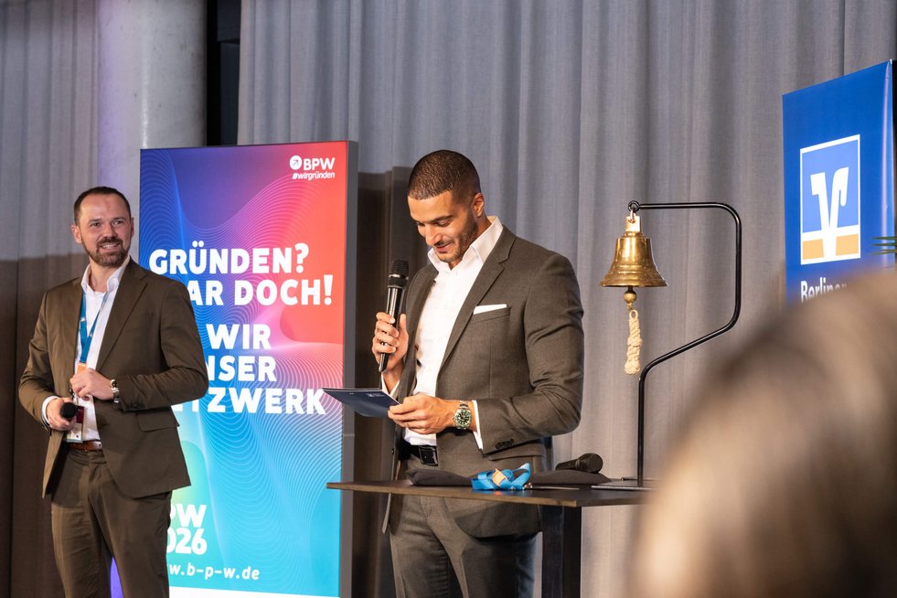 Zwei männliche Personen stehen auf einer Bühne und halten eine Rede