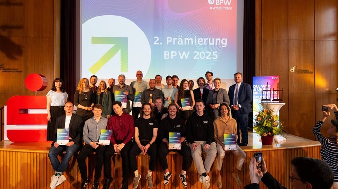 Gruppenfoto mit vielen Menschen, die entweder stehend oder auf einer Bühne sitzend Richtung Fotograf schauen