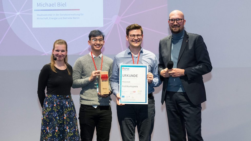 Preisverleihung. Zwei Sieger werden von der Moderatorin und dem Laudator umrahmt für ein Gruppenfoto