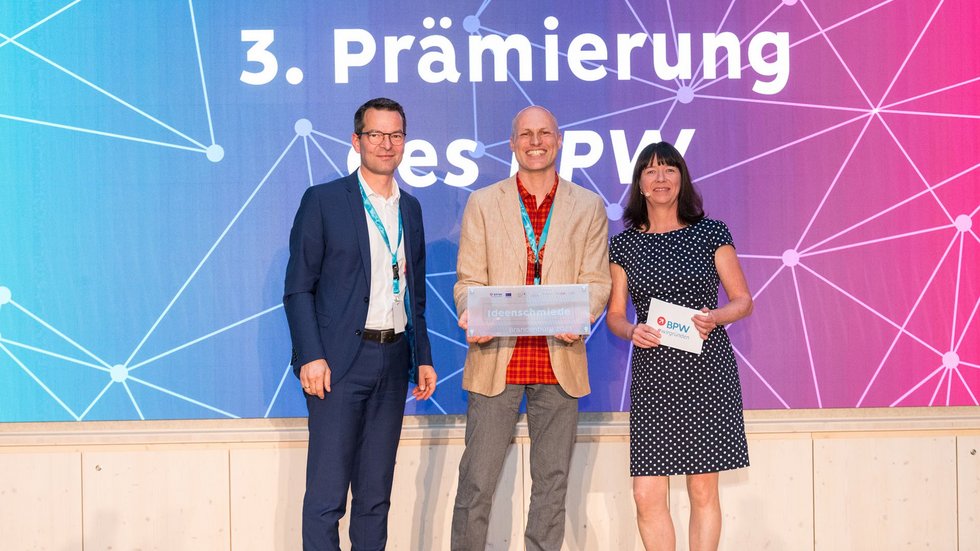 Auszeichnung auf einer Bühne, drei Personen blicken Richtung Publikum und halten eine Plakette