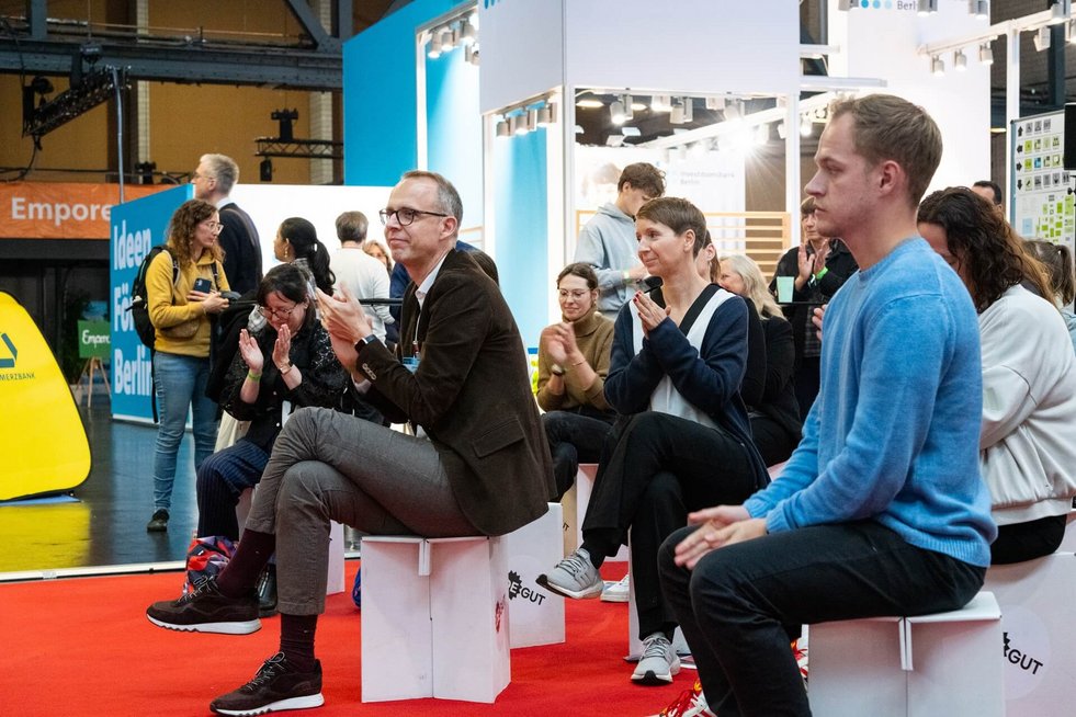 Blick ins Publikum auf dem Marktplatz der Messe deGUT. Gäste sitzen interessiert auf Hockern und blicken zu den Personen auf der Bühne.