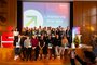 Große Gruppe steht und sitzt auf einer Bühne, alle blicken für ein Gruppenfoto in Richtung eines Fotografen