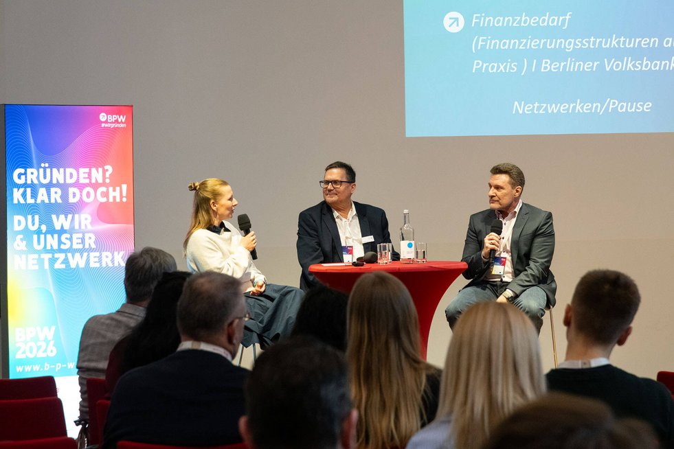 Drei Personen sprechen auf einer Bühne vor Publikum.