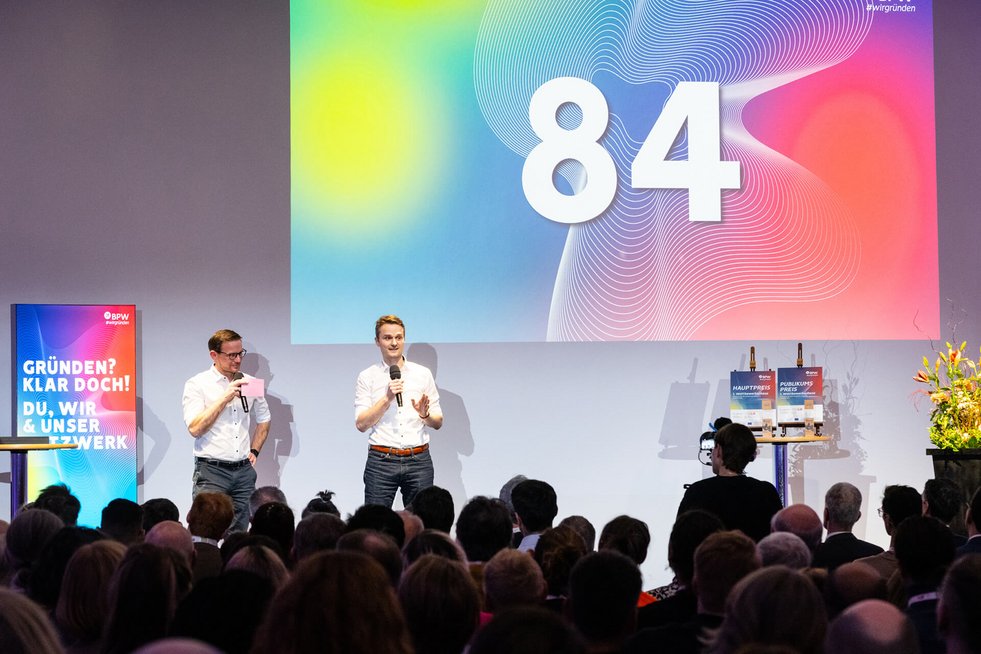 Zwei Männer sprechen mit Mikrofonen auf einer Bühne vor einem großen Publikum. Im Hintergrund ist eine Projektion mit der Zahl „84“ zu sehen.