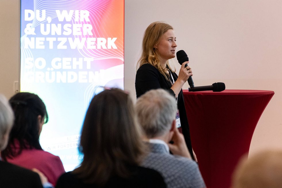 Eine Frau steht auf einer Bühne und spricht mit einem Mikrofon zu einem Publikum