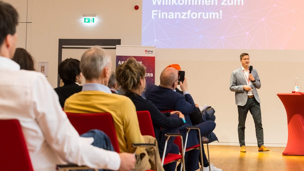 Referent spricht ins Mikrofon zu den sitzenden Teilnehmer:innen 