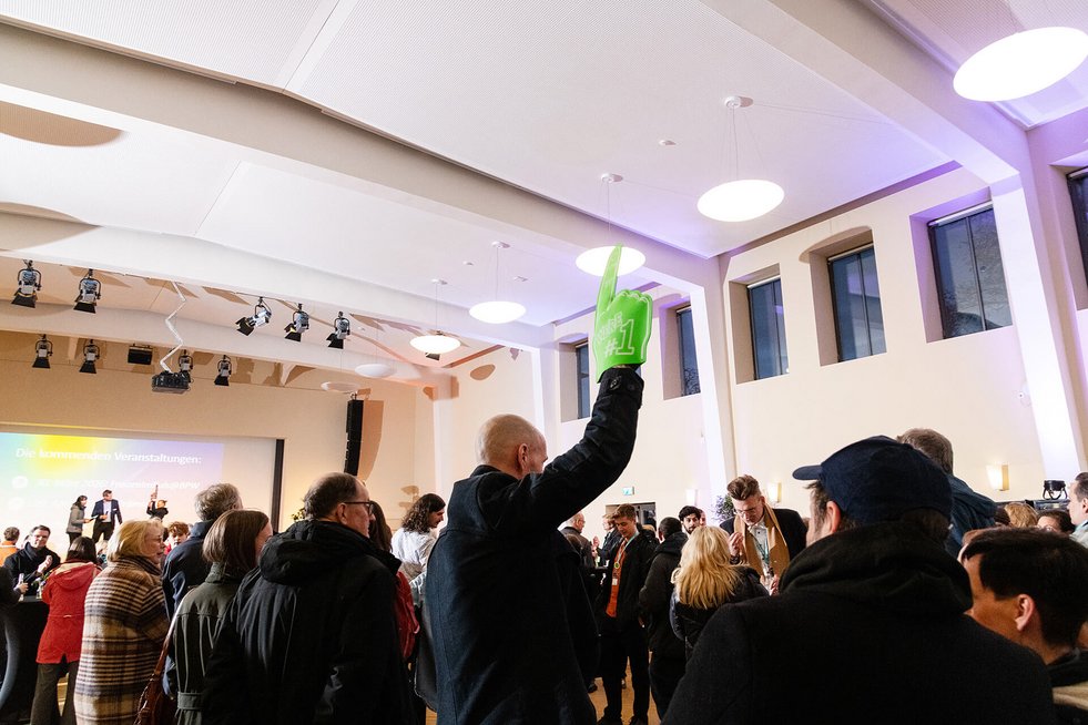 Menschen stehen in einem Veranstaltungssaal, eine Person hält eine grüne Schaumstoffhand in die Höhe.