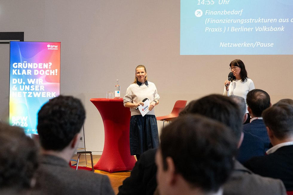 Zwei Moderatorinnen sprechen auf einer Bühne vor Publikum.