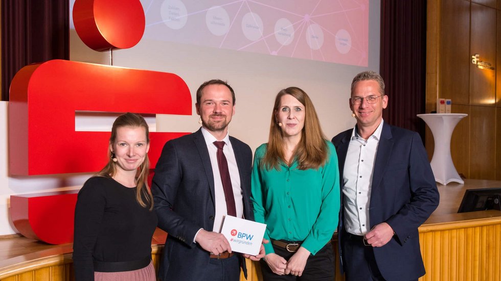 Vier Personen stehen vor einer Bühne für ein Gruppenfoto zusammen.