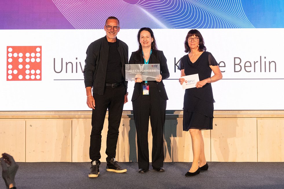 Drei Personen stehen auf einer Bühne bei der BPW-Prämierung. Die Person in der Mitte hält die Auszeichnung „Ideenschmiede Berlin 2026“, daneben steht eine Moderatorin mit einer BPW-Karte. Im Hintergrund ist das Logo der Universität der Künste Berlin zu sehen.