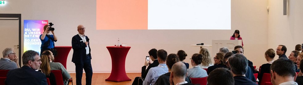 Blick in einen Veranstaltungssaal, in dem ein Referent auf einer Bühne zu einem Publikum spricht