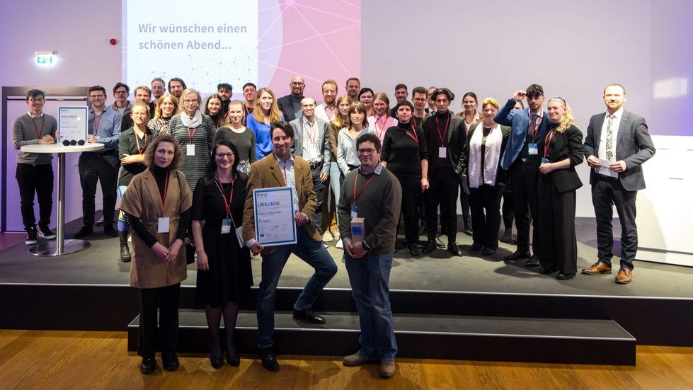 Gruppenbild auf einer Bühne. Die Laudtoren und Finalist:innen der ersten Phase stehen zusammen.