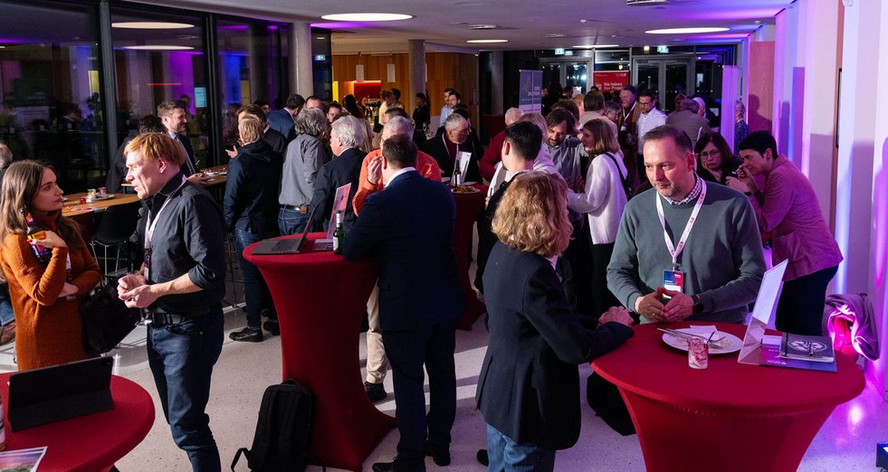 Zahlreiche Personen stehen bei einer Networking-Veranstaltung in einem hell beleuchteten Innenraum. Sie unterhalten sich in kleinen Gruppen an Stehtischen. Die Atmosphäre wirkt lebendig und kommunikativ.
