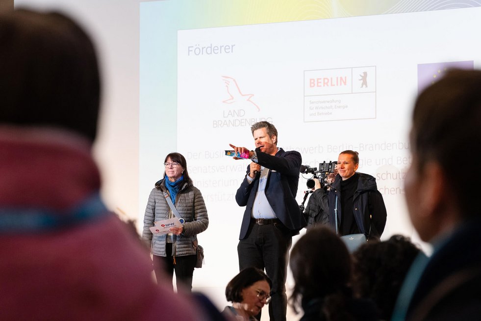 Mann spricht auf einer Bühne in ein Mikrofon, zwei Frauen stehen neben ihm, Publikum im Vordergrund.