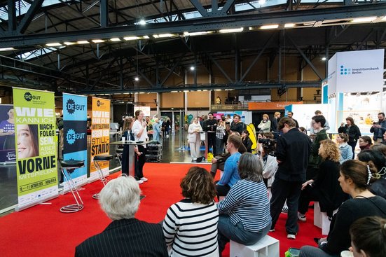 Marktplatz auf der Messe deGUT, die Hocker sind alle besetzt mit Gästen, die die Veranstaltung verfolgen. 