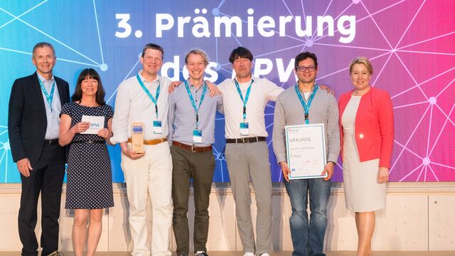 Sieben Personen stehen auf einer Bühne für ein Gruppenfoto und halten eine Urkunde ins Bild